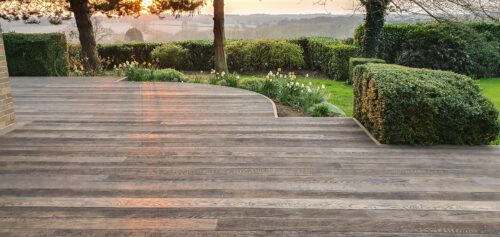 Millboard Weathered Oak composite decking with a naturally aged timber look, installed in a garden setting with sculpted hedges and a sunset background.